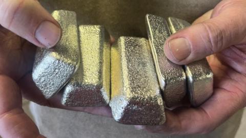 A close shot of five shiny whitish-yellow Cornish tin ingots held in a mans hands.