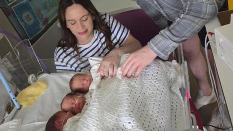 A photo of a woman looking down at three babies who are sleeping. She has got a white top on with blue stripes. 