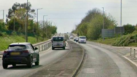 A main road with a number of vehicles driving towards a flyover. There are cycle lanes on each side of the road which is lined with trees.