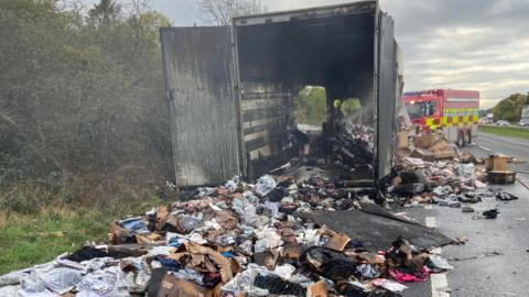 A burnt out lorry with its back doors open on the A1. Items spill out from the lorry onto the road. A fire engine is in the background.