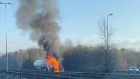 A picture of the lorry on fire from the opposite side of the carriageway. The carriage of the lorry is engulfed in fire, with black smoke coming off it into the air.