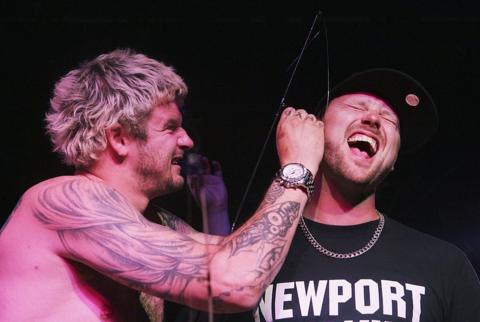 Dirty Sanchez star Mathew Pritchard (left) photographed piercing the ear of his fellow TV star, Lee Dainton (right). Pritchard has shaggy blonde hair with is slightly grown out at the root. He is topless, has arm tattoos and holds his right hand up to Dainton's ear. Dainton holds his mouth wide open in pain and wears a black cap and black t-shirt which reads 'newport'. He wears a gold chain and has a short stubbled brown beard.