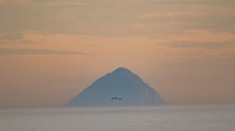 Ailsa Craig in the distance in a deep orange sunset and still sea. Three birds fly in front of it.
