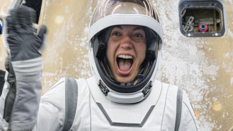 An astronaut waves at the crowd. She's smiling broadly and wearing her white-and-grey spacesuit and is standing infront of the Dragon capsule.