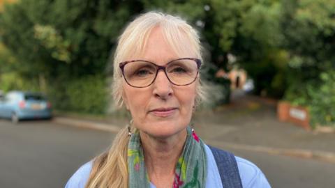 A woman with  greyish and blonde hair in a side ponytail and glasses. She is wearing a dark blue waist coat and light blue shirt and a green scarf