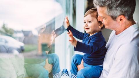 A smiling toddler boy - wearing a blue top, jeans and stripy socks - presses his hand against a window which looks out onto a residential street. He is being held by a man in a long-sleeved white top, with short black grey hair.