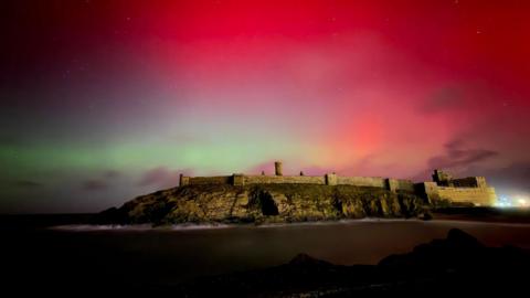 The ruins of Peel Castle on an islet, with a green strip of light above it under a bright pink and red sky dappled with stars.