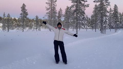 A woman in black trousers, a white ski-style jacket, a black beanie hat and black gloves stands on snow-covered ground in front of snow-covered trees.