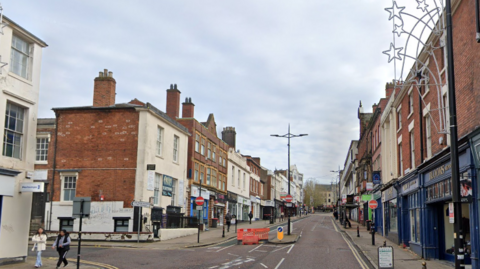 The street has shops on either side. A barrier on the left hand side of the street prevents traffic from driving on to it.