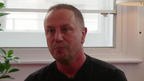 A man in a black shirt sits in front of a white-framed window, looking serious. A leafy plant and a lit lamp are partially visible at the edges of the image.