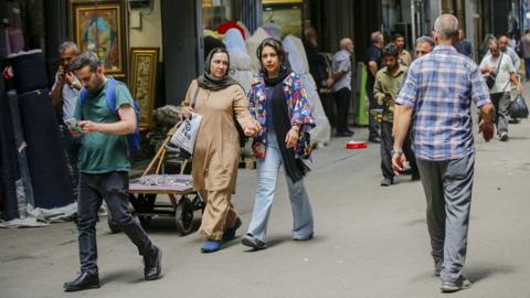 Several people walk on the streets of Tehran on a shaded street in July.