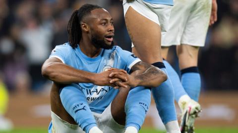 Antoine Semenyo in light blue Manchester City jersey, white shorts and light blue socks sits on the pitch during a Premier League game. He has his knees raised up towards his chest and is resting both arms across them, with his hands clasped together. He has a frustrated expression on his face as he looks towards the right of the image. The legs of two City team-mates can be seen behind him