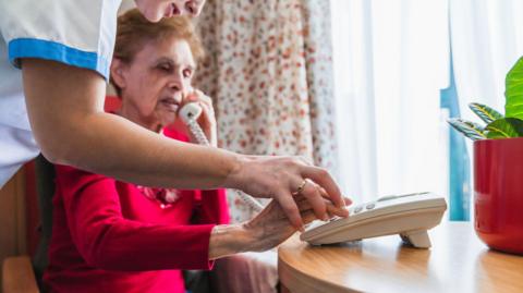 A nurse in blue and white uniform helps and elderly lady in a red jumper type numbers into a phone while sitting at a wooden desk