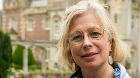 A woman with short, blonde hair and wire-rimmed glasses in a light blue jumper with a blue striped blouse underneath stands in front of Abbotsford House the former home of Sir Walter Scott