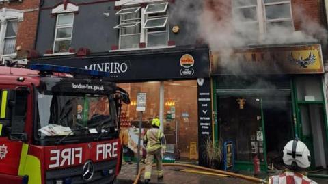 Scene shows fire engine in front of burning building on high street. Smoke can be seen billowing from the building and fire officers.