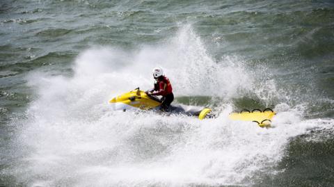 A person in RNLI uniform on a jet ski towing an empty stretcher. The sea is quite choppy and the jet ski has created a lot of wash.