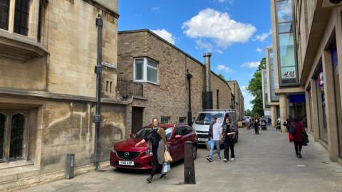 Image taken from Christ's lane, showing shops on one side, pedestrians in the foreground and brick buildings in the background.