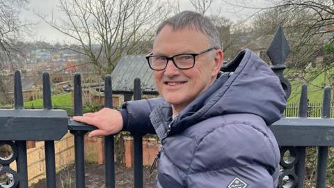 Mike Stonard is standing next to a set of railings. He is looking directly at the camera and smiling. He is wearing a black padded jacket.