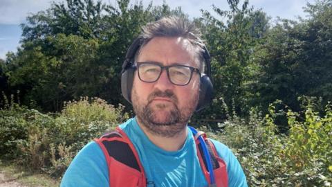 A man wearing a light blue t-shirt with red straps over each shoulder. He has shirt brown hair and facial hair, thick black glasses and black headphone. He is looking into the camera and is stood in front of green hedgerow