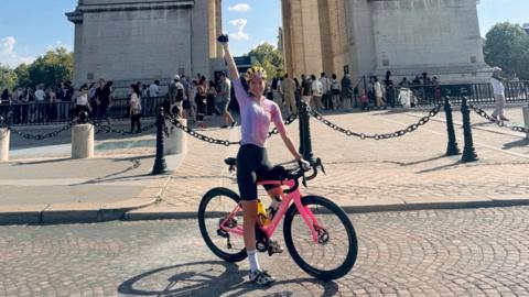 Amy Hudson at the Arc de Triomphe in Paris. She is dressed in a pink and white cycling top, black shorts and a yellow helmet. She is sitting on her pink bike with her arm pointing in the air, with the base of the Arc de Triomphe behind her in light coloured stone.