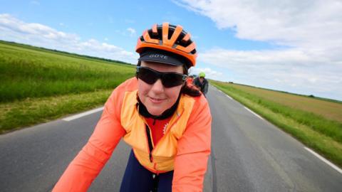A woman wearing sunglasses, a bright orange jacket and an orange cycle helmet is riding a bike towards the camera along a road which runs between two fields and under blue skies with a few clouds. Behind her are more cyclists.