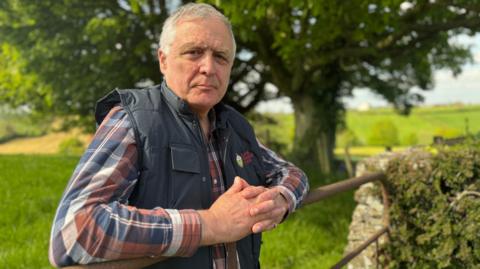 President of the Ulster Farmers' Union William Irvine leaning on a farm gate on a sunny day.  He has short, grey hair and is wearing a red, white and blue checked shirt under a navy bodywarmer.  There are trees and open fields behind him. 