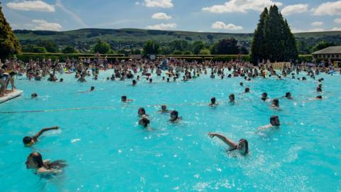 An outdoor swimming pool filled with dozens of people in the water swimming. Hills can be seen in the background.