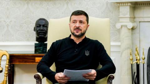 Zelensky wearing a black long-sleeved t-shirt and holding some A4 paper, has a frowning expression as he sits in a white armchair in the White House next to Biden at a news conference. Behind him is a fireplace and cast iron bust