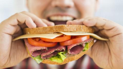 A man holds up a sandwich to his mouth. The sandwich has cheese, tomato, pastrami and lettuce in it