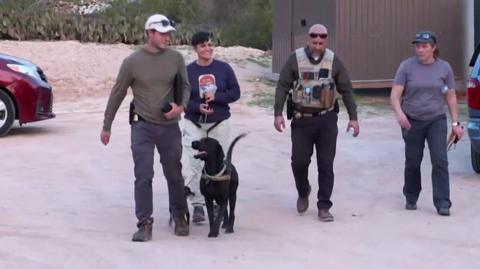 A group of four people walk with a black sniffer dog on a lead outdoors in Tuscon.