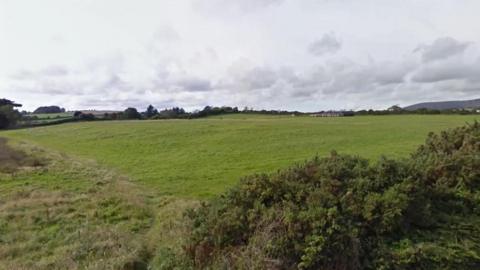 Large green field in Braddan edged by hedgerows. The sky above is pale in colour with fluffy clouds.