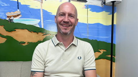 David Shaw sits in front of a mural depicting the Norfolk coast in yellow, green and blue. He is wearing a cream polo shirt. He is bald and has grey stubble.