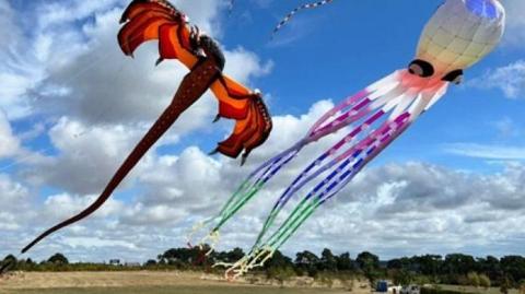 A red dragon and white octopus kits flying over a large flat open field with trees on the horizon