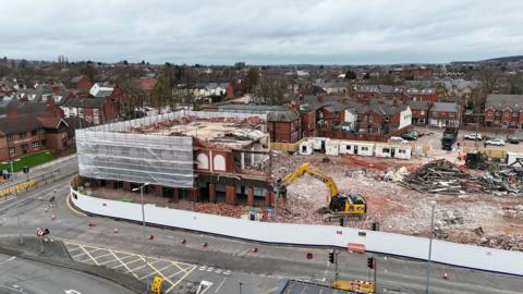Drone shot of a building site where a two story brick buildings has been extensively demolished by a digger and much of the rest of the site has been cleared
