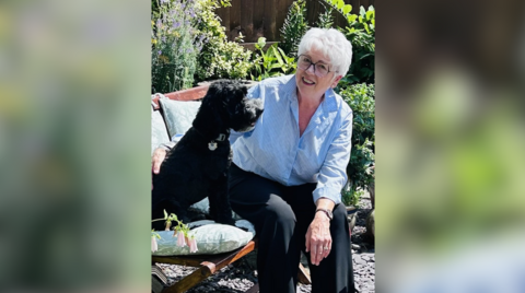 A woman in a blue and white shirt, black glasses and light hair, sitting on a bench with a labradoodle-style dog with a dark coat.