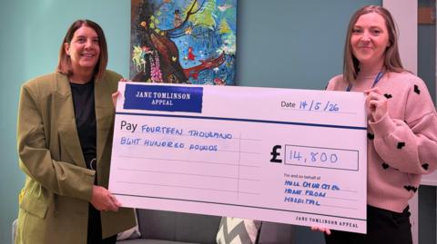 Two people are standing indoors against a blue-green wall, holding a large ceremonial cheque from the “Jane Tomlinson Appeal.” The cheque is dated “14/5/26” and made out for “Fourteen thousand eight hundred pounds,” with the amount “£14,800” written in a large box on the right. Beneath the amount, handwritten text reads “For and on behalf of Hull and Ousea Home From Hospital” 