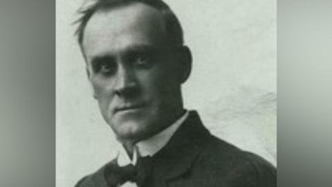 A black and white image of David Danskin. A clean-shaven man with short black hair is wearing a black bow-tie, white shirt and a black blazer