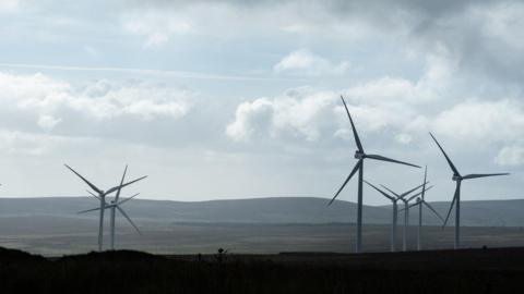A view of a wind farm from Benbradagh Summit in County Londonderry on a cloudy day.  There are seven white turbines with three spokes each in a field.  There are rolling hills on the horizon. 