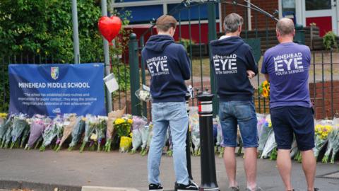 Three men stood with jumper's reading 'Minehead Eye' have their backs to the camera, looking at a row of flowers which have been laid in front of a fence. A sign saying 'Minehead Middle School' is attached to the railings.