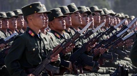 Korean People's Army soldiers march during a mass rally on Kim Il Sung square in Pyongyang. They are seen marching in a line, each staring forward holding a rifle at a 45-degree angle. The rifles have bayonets on the end. 