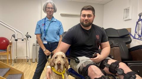 A man sits in a mobility chair with his legs at a 90' angle to his body. He is wearing a black t-shirt and his legs are supported with black harnesses. He as short dark hair, a dark beard and cream shorts. Sat beside him is a blonde English Setter dog wearing a yellow necktie. The dogs lead is held by a woman who stands behind the pair. She is wearing a blue polo shirt with logos, dark jeans, glasses and short grey hair. Hospital therapy equipment can be seen behind the group. The walls are cream and the flooring is a light wood laminate.