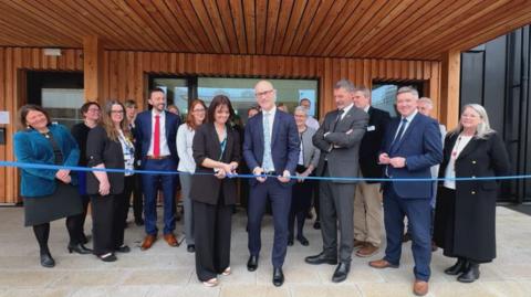 A group of men and women standing outside, with one man in a suit cutting a blue ribbon to a building. 
