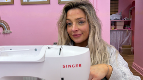 Shelly sits at a table with a sewing machine in front of her. The walls behind her are painted pink. She has long, blonde hair and is wearing a grey sweat top.