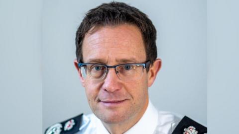 Pictured is Chris Noble against a pale blue background. He is slightly smiling, wearing rectangle frame glassed and has brown hair and blue eyes. He is wearing a white shirt with black shoulder trim and police badges. 