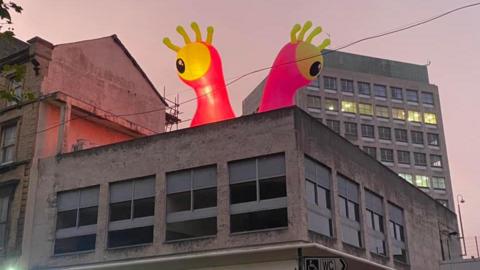 A two-eyed red inflatable monster with boggly eyes looming over a building in Wolverhampton. It is casting a red hue in the sky and there is another high-rise building in the background. 