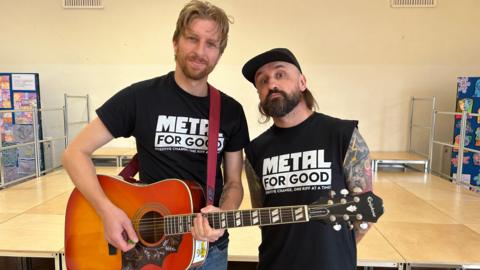 Baz Mills and Adam Thistlethwaite,  who is holding a guitar, are standing in the middle of a hall at Trumacar Community Primary School. They are both wearing black T-shirts which feature the message "Metal for Good". 