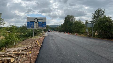 A newly resurfaced road under a cloudy sky. The road surface is dark grey, and on one side is a sign showing that the M5 motorway junction is ahead, with white text on a blue background. There is plant and tree debris on one side of the road and cones either side in the distance