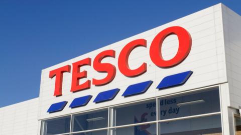 A Tesco logo on a white building. There is a blue sky above. 