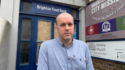 A bald man in a pale blue shirt, standing in front of a navy blue and glass door which has the words Brighton Food Bank on it in white