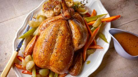 A roast chicken surrounded by sliced carrot and celery on a large white platter with a jug of gravy
